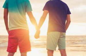 Happy gay couple holding hands watching sunset on the beach